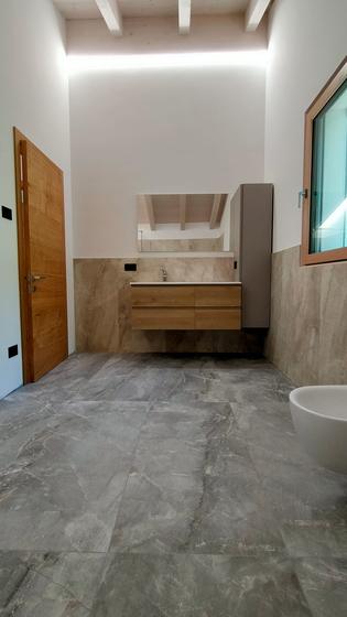 Bagno con pavimento in gres effetto marmo grigio, mobile sospeso in legno, colonna e vasca sotto un soffitto con travi a vista
