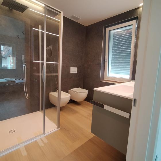 Bagno con rivestimento in gres scuro, pavimento effetto rovere chiaro, box doccia in cristallo e mobile lavabo sospeso