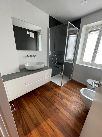 Bagno con pavimento in legno scuro, mobile bianco con top grigio e lavabo da appoggio, box doccia con rivestimento nero