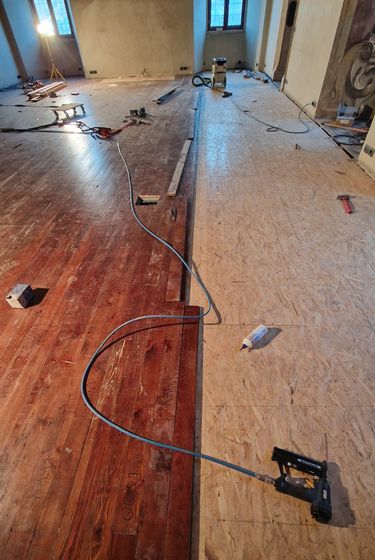 Sala in cantiere: sopra il vecchio pavimento in tavole di legno si stende un nuovo sottofondo in pannelli, con sparachiodi e attrezzi appoggiati a terra