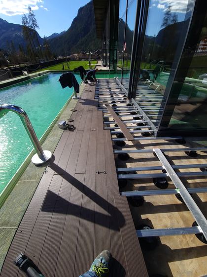 Fascia di decking posata lungo il lato lungo della vasca mentre la sottostruttura prosegue verso la vetrata, con un posatore al lavoro sul fondo