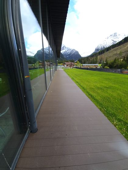 Camminamento in decking posato lungo la vetrata, con il bordo netto sul prato e le montagne sul fondo