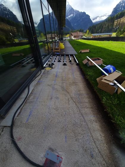 Sottostruttura in corso lungo la vetrata, con un tratto di decking già chiuso in fondo e le scatole dei supporti appoggiate sul prato