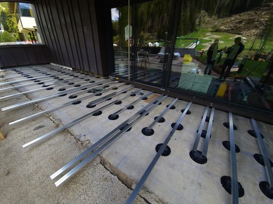 Travetti metallici appoggiati su supporti regolabili e posizionati secondo la tracciatura blu, davanti alla facciata vetrata con parete in lamiera scura