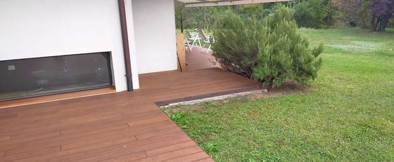 Terrazza in decking a doghe che affianca la casa e si chiude con un taglio netto sul prato, accanto a un grande cespuglio di rosmarino