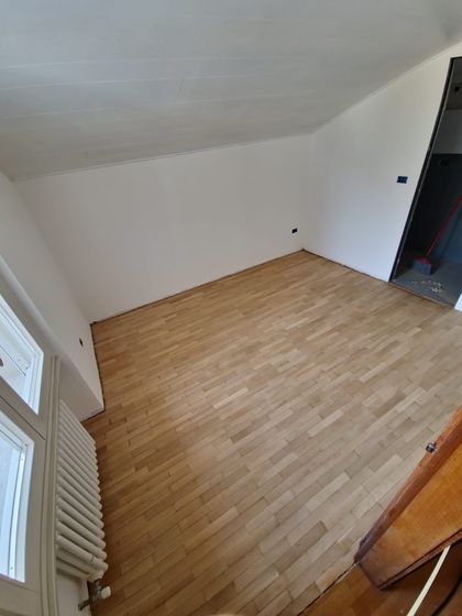 Stanza mansardata vuota con pavimento in legno a listoncini appena levigato, chiaro e opaco, e radiatore sotto la finestra
