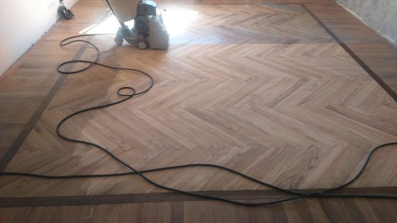 Levigatrice a nastro al lavoro su un pavimento in legno a spina di pesce, con il cavo steso sulla superficie già passata
