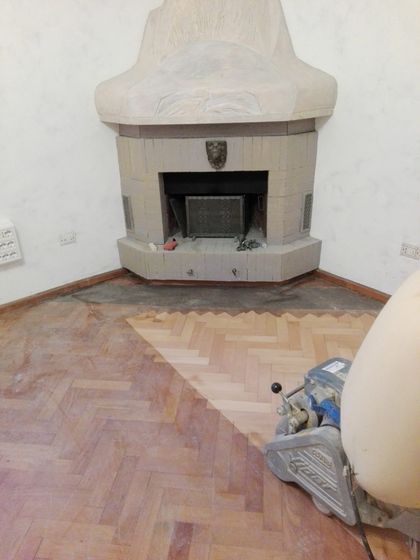 Stanza con camino in muratura ad angolo e cappa che sale al soffitto: il pavimento in legno a spina di pesce è levigato solo in una fascia, il resto è ancora scuro, con la levigatrice in campo
