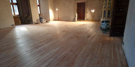 Grande sala di un edificio storico con il pavimento in tavole di legno larghe appena levigate; a bordo campo la levigatrice a nastro, due aspiratori e i fari da cantiere accesi