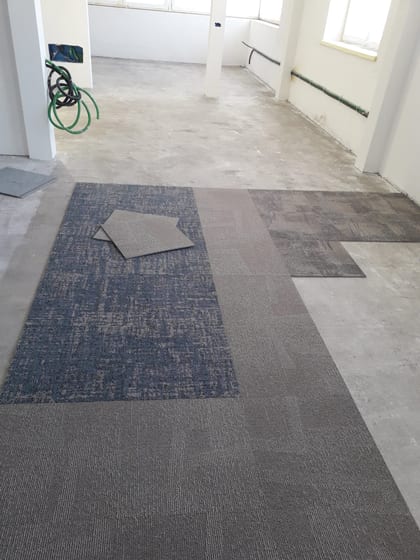 Posa di moquette in un ambiente in cantiere: un campo blu-grigio e campi grigi accostati sul fondo ancora scoperto, con due pezzi appoggiati sopra