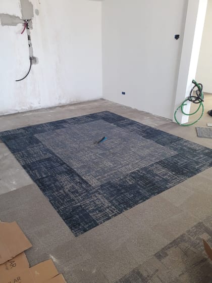 Stanza in cantiere vista d'angolo: riquadro di moquette blu-grigia posato a filo del campo beige, cartoni e tubi a bordo campo, fondo ancora scoperto lungo le pareti