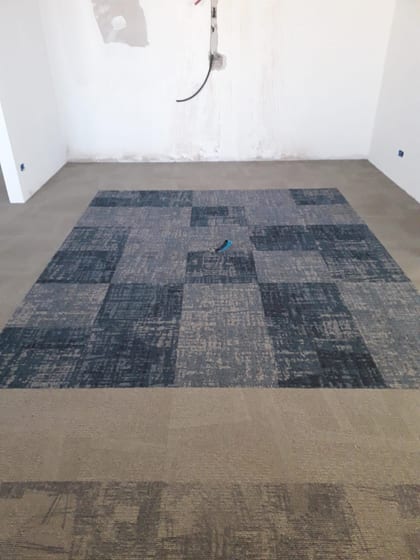 Riquadro di moquette con cornice blu-grigia attorno a un campo più chiaro, posato dentro la moquette beige di una stanza ancora da finire, con un attrezzo appoggiato sopra