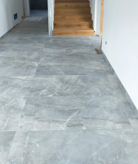 Corridoio con pavimento in grandi lastre grigie venate e scala rivestita in legno chiaro, muri ancora grezzi